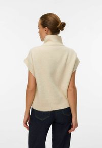 Maglione corto a maniche corte di colore crema con colletto alto e orlo a coste. Tessuto morbido, vestibilità rilassata, indossato sopra pantaloni in denim scuri.