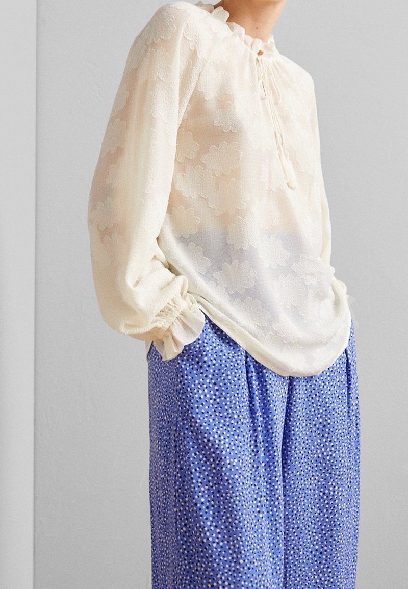 Blouse crème à manches longues avec texture florale et poignets volantés, associée à un pantalon bleu taille haute avec petits pois noirs et blancs.