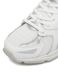 Baltas ir sidabro spalvos sportinis kedas iš arti, rodantis akių tinklo audinį, siuvines viršutines dalis, raištelius ir tekstūruotą gumos pado paviršių.