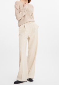Femme portant un pull beige texturé, un pantalon large crème taille haute et des chaussures en cuir noir, debout devant un fond blanc.
