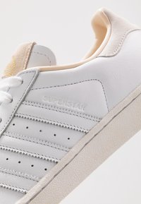 Zapatilla blanca de cuero con suela de goma texturizada, presenta perforaciones, tres rayas laterales y una pestaña de talón de gamuza beige. "SUPERSTAR" grabado en el costado.