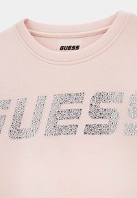 Camiseta rosa claro con cuello redondo, con grandes rhinestones plateados formando la palabra "GUESS". Textura de tejido suave.