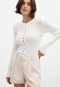 Valkoinen ribattu pitkähihainen paita, jossa on pyöreä kaula-aukko ja etunapit. Yhdistetty vaaleanbeigeihin korkeavyötäröisiin shortsit, joissa on sivupleatit.