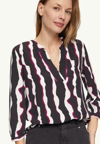 Zwarte blouse met witte en magenta golvende strepen, met een knoopsluiting en driekwart mouwen. Gladde stofstructuur.