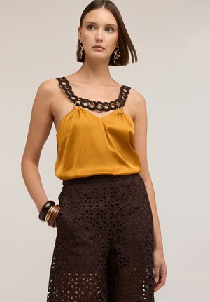 Kobieta w satynowym musztardowożółtym topie na ramiączkach z czarnym pętlowym paskiem oraz ciemnobrązowych szortach z kwiatowej koronki, uzupełniona bransoletkami i kolczykami.