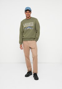 Felpa verde oliva con logo "LACOSTE" e grafica di montagna, pantaloni beige e sneakers nere, abbinata a un cappellino azzurro chiaro.