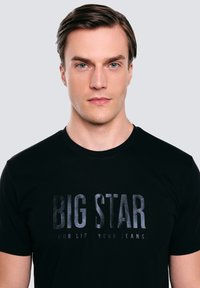 Černé bavlněné tričko s velkým grafickým potiskem v tmavě šedé, na kterém je napsáno "BIG STAR" a "OUR LIFE YOUR JEANS." Krátké rukávy a kulatý výstřih.