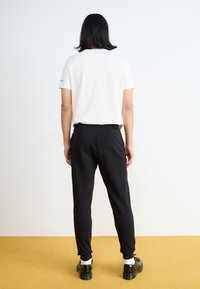 Sorte joggebukser med elastiske manchetter, parret med en ensfarvet hvid t-shirt. Modellen har sorte sko på med hvide sokker foran en gul baggrund.