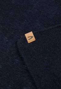 Mørkeblå strikket stof med et tekstureret mønster, der har et lille tan lædermærke med et logo syet ind i hjørnet.