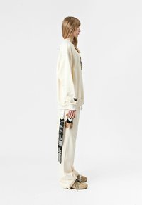 Sudadera y pantalones color crema; detalle notable de oso de peluche en los pantalones y cinta negra con texto. Modelo sosteniendo un oso de peluche en la mano.