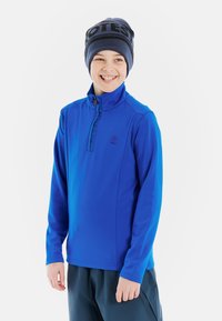 Pullover blu a maniche lunghe con colletto alto, chiusura con zip e un piccolo logo sul lato sinistro del petto, abbinato a pantaloni scuri e indossato con un berretto.