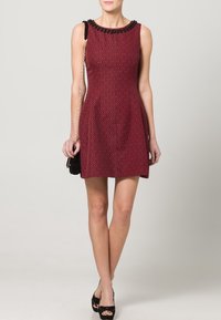 Robe sans manches rouge avec un motif géométrique, encolure ornée de perles noires, silhouette évasée, associée à un petit sac à main noir et des talons noirs.