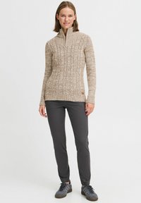 Beiger Strickpullover mit einem Viertelreißverschluss, strukturiertem Design und langen Ärmeln; kombiniert mit dunkelgrauen Hosen und grauen Schuhen.