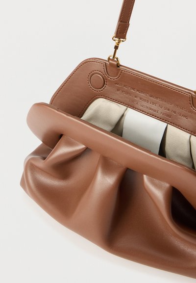 Καφέ δερμάτινη τσάντα χειρός με λεπτομέρειες ραφής, χρυσά μεταλλικά στοιχεία και μπεζ υφασμάτινη επένδυση, με ανάγλυφο κείμενο μέσα στο πλαίσιο.