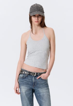 Jeune femme portant un haut dos nu gris, un jean bleu avec une ceinture cloutée et une casquette de baseball grise usée, debout devant un fond uni.