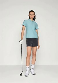 Šviesiai mėlynas polo marškinėliai su apykakle ir trimis mygtukais, derinami su tamsiomis šortais. Figūra laiko golfo lazdelę, vilkėdamas baltus sportinius batus ir pirštines.