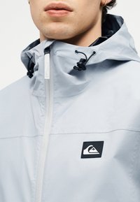 Κοντινό πλάνο ενός ατόμου που φοράει ανοιχτό γκρι μπουφάν με φερμουάρ και κουκούλα, με μαύρο λογότυπο στο στήθος και ρυθμιζόμενα κορδόνια.