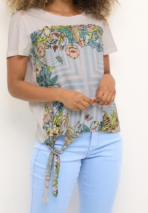 Mujer que lleva una camiseta beige de manga corta con estampado colorido de flores y pájaros, con un nudo atado a un lado, combinada con pantalones azul claro.