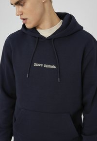 Hoodie bleu marine en tissu doux, mettant en avant un logo brodé "KEITH HARING" en blanc et jaune sur la poitrine et une poche avant.