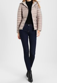 Kvinde iført en beige puffet jakke, sort turtleneck, mørke skinny jeans og sorte ankelstøvler, der står foran en helt hvid baggrund.