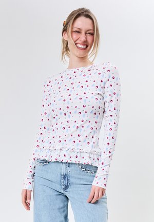 Smilende kvinde iført en hvid langærmet top med røde og blå prikker, kombineret med lyseblå jeans med bølget denimkant.