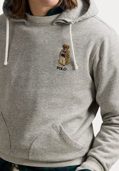 Haut à capuche gris avec des cordons blancs ; présente un ours brodé portant une chemise "Polo" bleue. Dispose d'une poche frontale et de poignets côtelés.