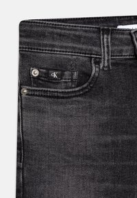 Svart denimjeans med subtil textur, silverfärgade knappar och en märkt etikett med "cK" på framfickan.