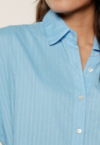 Jasnoniebieska koszula zapinana na guziki z pionowym wzorem w paski, z kołnierzykiem i gładką bawełnianą teksturą. Z przodu znajdują się białe guziki.