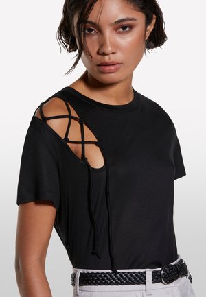 T-shirt noir avec un design à épaules dénudées, en tissu noir entrecroisé, à manches courtes et d'une texture lisse. Porté avec une ceinture noire tressée.