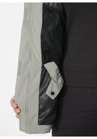 Leichtgewichtige graue Jacke mit Ärmeln, die ein strukturiertes schwarzes Mesh-Panel und eine Druckknopftasche aufweisen. Glattes Material mit kontrastierenden Akzenten.