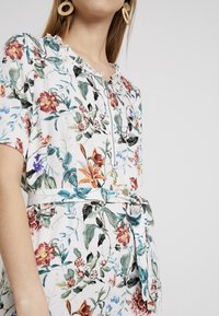Mujer con un vestido floral blanco de mangas cortas, una cremallera frontal, un cinturón de tela atado en la cintura y grandes pendientes circulares de mimbre.