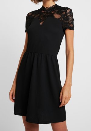 Czarna sukienka z krótkimi koronkowymi rękawami, wysokim dekoltem i dopasowanym gorsetem przechodzącym w rozkloszowaną spódnicę. Z ozdobami z kwiatowej koronki.
