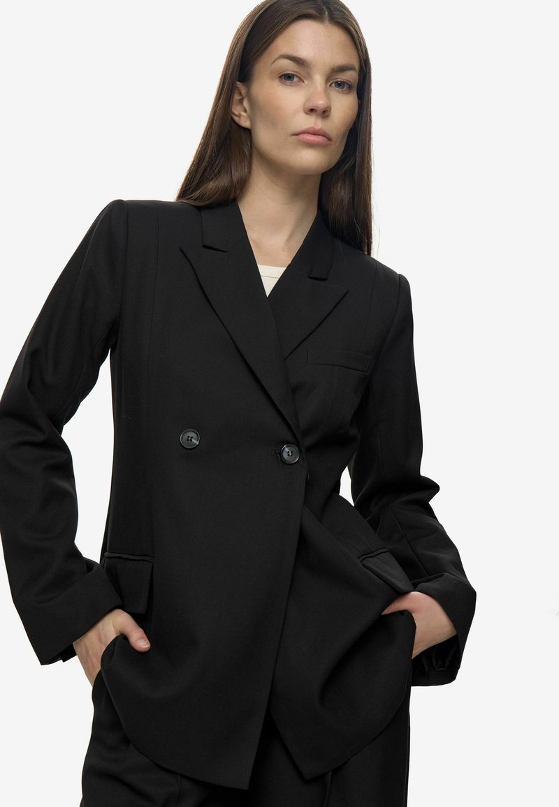 Zwarte double-breasted blazer met een geknipt revers, twee voorzakken en zichtbare stofstructuur. Grote zwarte knopen als hardwareaccenten.