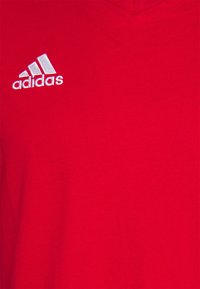 Punainen puuvillainen t-paita, jossa on V-pääntie. Vasemmassa rintakehässä on valkoinen brodeerattu Adidas-logo. Sujuva pinta.