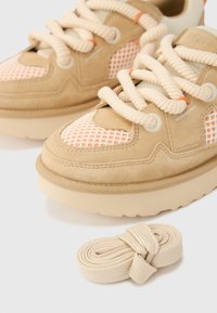 Baskets beiges avec des panneaux en mesh orange clair, des lacets épais crème, une semelle en caoutchouc et un marquage discret. Un lacet beige supplémentaire roulé est inclus.