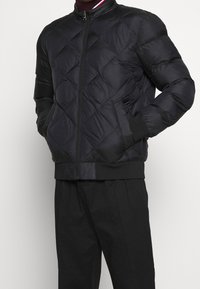 Giacca imbottita nera trapuntata realizzata in materiale sintetico, con zip frontale, polsini a coste e design con collo rotondo.