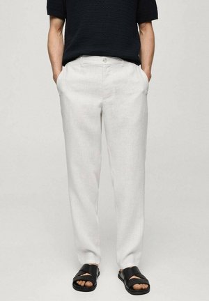 Persoon die witte linnen broek, zwart topje met korte mouwen en zwarte sandalen met kruisbandjes draagt, staand met handen in de zakken.