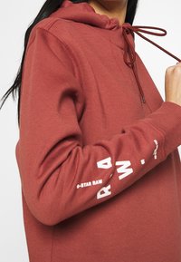 Mikina barvy rezavé červené s tkaničkou, žebrovanými manžetami a nápisem "G-Star Raw" na rukávu. Měkká, strukturovaná tkanina a volný střih.