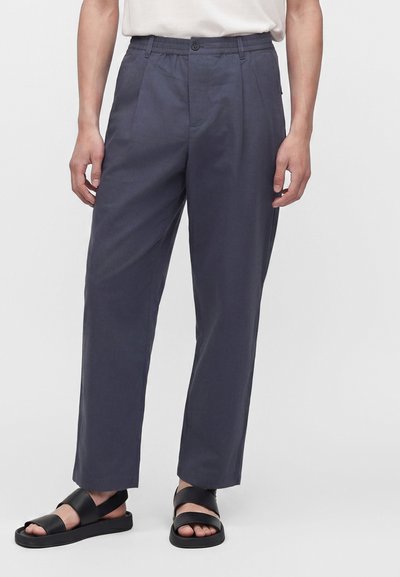 Homme portant un pantalon fuselé bleu marine associé à des sandales en cuir noir et une chemise blanche rentrée.