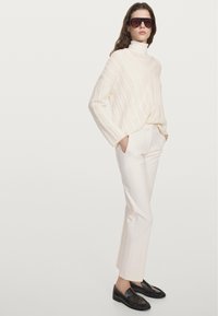 Gebreide crème kabeltrui met hoge hals, gecombineerd met op maat gemaakte witte broek en zwarte loafers. Model draagt een donkere zonnebril.