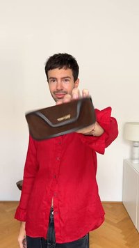 Estuche de gafas rectangular marrón con borde cosido y logo dorado, sujetado contra un fondo liso por una persona con una camisa roja.