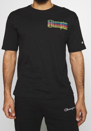 Mand iført sort Champion t-shirt med farverigt gentaget logo på brystet og matchende sorte bukser med Champion-logo på låret.