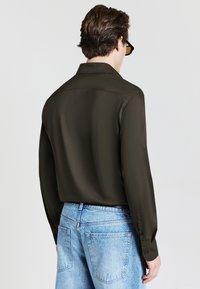 Camicia a maniche lunghe verde oliva con yoke sul retro, abbinata a jeans in denim azzurro chiaro. Tessuto con texture liscia, colletto classico e polsini con bottoni.