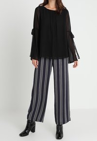 Svart genomskinlig blus med veck och volangärmar, tillsammans med bredrandiga byxor i marinblått och vitt. Glansiga svarta ankelskor fulländar looken.