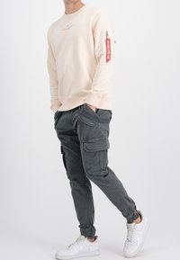 Cremfarbener Sweatshirt mit Text auf der Vorderseite und rotem Reißverschluss-Taschen-Detail. Graue Cargohosen mit mehreren Taschen. Weiße Sneaker.