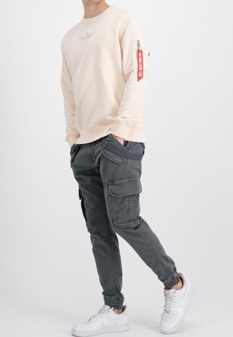 Cremfarbener Sweatshirt mit Text auf der Vorderseite und rotem Reißverschluss-Taschen-Detail. Graue Cargohosen mit mehreren Taschen. Weiße Sneaker.