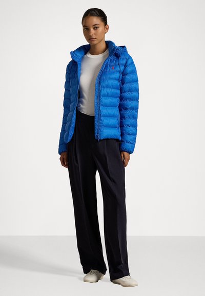 Blauwe pufferjack met capuchon, met een geweven textuur, voorzijde met rits en klein logo. Draag een witte shirt met losse zwarte broek en witte schoenen.