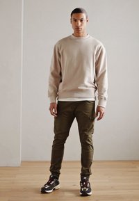 Beżowa bluza z okrągłym dekoltem, oliwkowe spodnie cargo i czarne sneakersy z akcentami. Gładka tekstura i luźny krój w casualowym stylu.