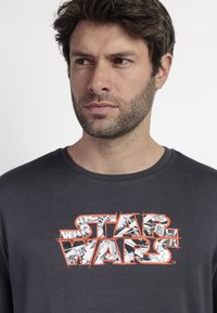 Camiseta de algodón de color gris oscuro con un estampado gráfico de "STAR WARS" en blanco y naranja, con un fondo de estilo cómic. Escote redondo.