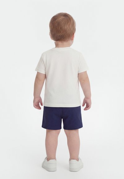 Παιδίατρος όρθιος με πλάτη προς τα εμπρός, φοράει λευκό κοντομάνικο μπλουζάκι, ναυτικά μπλε σορτς και λευκά αθλητικά παπούτσια σε απλό φόντο.